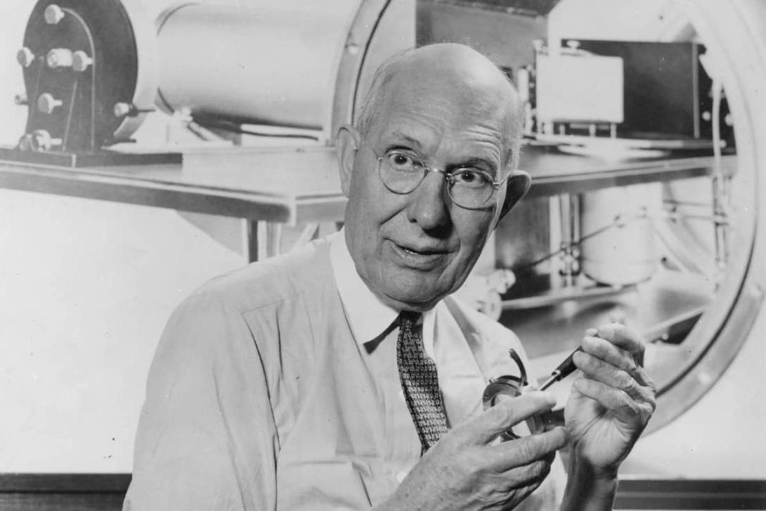 A black-and-white photo of Charles Kettering, sitting in a lab and holding machine parts.